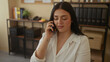 © Krakenimages.com - Woman talking on phone in office wearing white shirt with books and folders in background, conveying professional and confident atmosphere indoors.