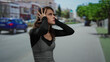 © Krakenimages.com - Woman standing outdoors on urban street covering ears appearing stressed, wearing a black top and checkered dress with a blurred cityscape in the background.