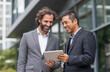 © 44designstore - Two professional businessman attire stands outside an office building, smiling and holding her iPad while looking at the screen