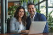 © 44designstore - Two smiling professional business man and woman, sitting together at a desk in a modern industrial-style office, collaborating and looking at a laptop screen