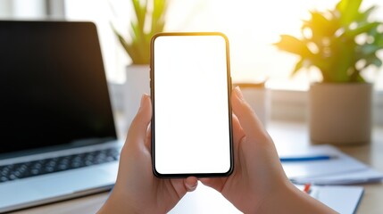 Poster - Hands holding a smartphone with a blank screen in a bright office setting with a laptop and potted plants in the background.
