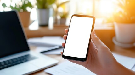 Poster - A person holds a smartphone with a blank screen in a bright, modern workspace featuring a laptop, documents, and potted plants in the background.