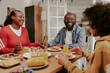 © AnnaStills - Black middle aged woman and Black middle aged man sitting at table smiling and talking with Black teenager while eating meal together, sharing food and enjoying family time indoors