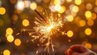 © rifqi - A hand holds a sparkler, creating a dazzling display of sparks against a backdrop of blurred golden lights.