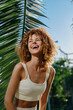 © SHOTPRIME STUDIO - smiling woman with curly hair wearing white crop top outdoors under sunny sky by tropical palm leaves, joyful and relaxed, vibrant summer atmosphere