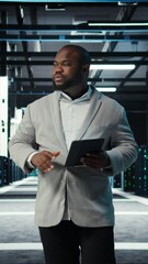 Poster - Vertical video IT professional looking over data center checkup checklist paperwork on tablet. African american man walking in server farm, reading documentation files on device, camera A