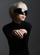 © SHOTPRIME STUDIO - Stylish woman in black turtleneck and oversized sunglasses, posing delicately with hands, evoking a sense of high fashion and modern elegance against a dark backdrop.