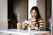 © Worawi - A young woman with glasses and a coffee cup uses her smartphone at a table, likely performing an online transaction or check.