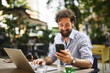 © Stockphotodirectors - Man sits at a table outside a cafe, using a laptop while looking at his smartphone. He smiles, sipping coffee, surrounded by greenery during a sunny day.