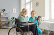 © Studio Romantic - Young friendly female nurse or social worker using modern tablet with senior woman with disability in wheelchair. Caregiver learning her elderly patient using digital gadget in nursing home.