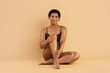 © Anastasiya - Smiling young Black woman in brown underwear sitting on floor in studio, hugging leg, posing confidently with glowing skin on beige background.