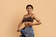 © Anastasiya - Excited young Black woman in brown underwear showing loose jeans after weight loss, smiling and pointing at her slim waist against beige background in studio.