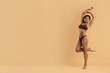 © Anastasiya - Happy young Black woman in underwear standing on one leg, arms raised playfully, smiling confidently against beige studio background, promoting healthy lifestyle and body joy.