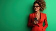 © svastix - A confident woman in a chic red suit holds a smartphone, smiling against a vibrant green background, showcasing modern fashion and empowerment.