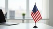 © 5 Grace Media - Small US desk flag on an office desk with a blurred laptop and window in the background
