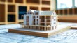 © sambath - A miniature model of a house with a wooden frame and white walls, placed on a blueprint with a wooden table in the foreground.