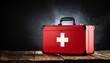 © Pastel - A red first aid kit with a white cross sits on a wooden surface against a dark background, symbolizing health and emergency preparedness