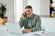 © insta_photos - Happy busy middle aged mature man senior entrepreneur talking on mobile phone looking at laptop using computer calling on cellphone working at home sitting at kitchen table.