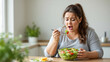 © Ventura - s a woman sitting at a table with a bowl of salad in front of her. She is holding a fork in her hand and appears to be eating the salad. On the left side of the table there is a plant