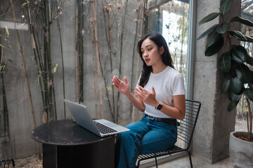 Aufkleber - Young Asian businesswoman gesturing while having a video conference on laptop in a modern office with bamboo plants