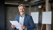 © Aryanedi - Confident mature businessman holding documents in modern office environment with professional smile