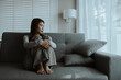 © Worawi - A young woman sits alone on a couch, holding her head in distress. The mood is somber, suggesting stress, anxiety, or emotional exhaustion at home.
