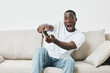 © SHOTPRIME STUDIO - Excited young Black man playing video games on a couch, showcasing joy and enthusiasm in a modern living room setting with light colors