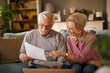 © Stockphotodirectors - A senior Caucasian couple, sitting on a sofa in their living room, are reviewing documents. The woman points at a paper, while the man holds other papers. They discuss financial matters.