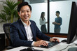 © Alex & M Studio - Friendly middle-aged asian businessman in a suit smiling at camera while working on a desktop computer at his desk in a modern office with colleagues in the background