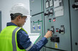 © JD Studio - Electrical engineer inspects control panel in substation room wearing safety helmet and reflective vest while checking equipment and recording data for maintenance and safety