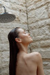 © SHOTPRIME STUDIO - Woman enjoying outdoor shower with wet hair and closed eyes, surrounded by textured stone wall, natural light highlighting serene and relaxed mood under water droplets.