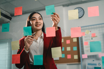 Poster - Businesswoman attaching colorful sticky notes to a glass wall, organizing tasks and ideas in a modern office environment