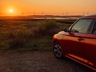 Wall Mural - A red car is parked in a field with wind turbines and a warm sunrise in the background. The car is facing the sun, and the sky is orange and pink. The scene is peaceful and serene. Early bird concept