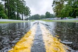 Close-up of a wet racetrack with double yellow lines during rainfall, creating ripples, onlookers in the background.