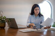 © Apichat - woman is sitting at a desk with a laptop and a stack of papers