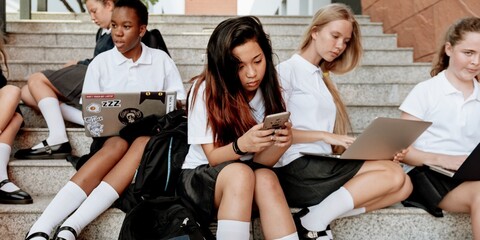 Poster - Group of diverse teenage girls in school uniforms sitting on steps. Girls using laptops and phones. School uniforms and technology in use by teenage girls. Diverse students relaxing at school stairs.