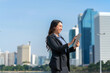 © StockPhotoRepublic - Smiling Asian businesswoman holding tablet with modern city skyline. Confident female professional outdoors in urban business district.