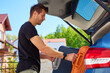 © Lazy_Bear - Man loading orange suitcase into car trunk on sunny day. Concept of preparing for summer travel, vacation or road trip with luggage