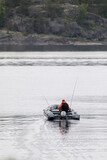 A lone fisherman in an inflatable motorboat heads out on a calm lake with fishing rods, surrounded by rocky forest shorelines.