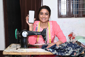 Happy traditional indian woman using sewing machine while showing smart phone with empty display advertisement, blank android phone screen.