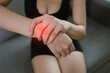 © Natthawadee - A woman sitting indoors holds her painful wrist with both hands, showing signs of discomfort, inflammation, or injury. Red glow indicates the affected area.