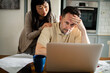 © Marko Geber - Worried couple reviewing finances together on laptop at kitchen table