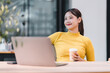 © PRIME STOCK LAB - Young Asian woman in yellow sweater sitting at desk with laptop, holding coffee cup, smiling and looking away, modern office background, relaxed and positive mood
