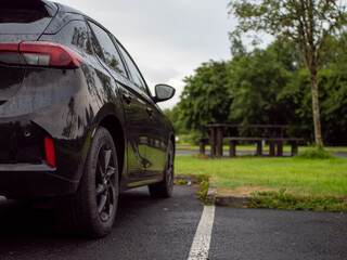 Wall Mural - Black family car parked in a parking lot of a park with barbecue or dining area, selective focus. Nobody. Car surface is with rain drops. Outdoor activity. Travel and tourism.