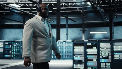 Poster - Happy manager listening music, walking in data center with blinking lights on rigs showing system activity. Cheerful man wearing headphones overseeing dimly lit server farm, camera A