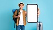 © photostockatinat - Happy traveler holding up a blank smartphone screen, ready for adventure. The traveler wears a hat, carries a backpack and stands near a suitcase, suggesting exploration.