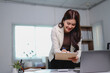 © MINAE - Young, smiling businesswoman standing at her desk, writing notes on a clipboard while working on a laptop in a bright, modern office environment, exuding confidence and professionalism