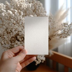 Poster - Hand holding blank white textured paper card with soft floral background and natural light