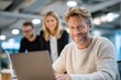 © The Little Hut - Confident Businessman Using Laptop Du Work Meeting with Colleagues in Modern Office Environment in the Background