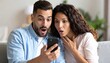 © M - Surprised couple reacting to unexpected news while looking at a smartphone in a modern living room